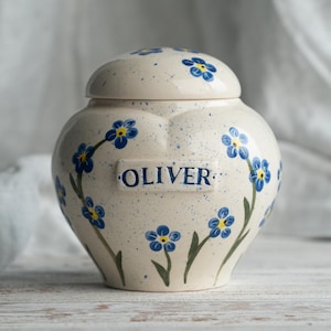 May include: Cream ceramic urn with a lid, decorated with blue forget-me-not flower designs and the name "OLIVER". The urn has a speckled finish and a rounded shape, suitable for memorial purposes.