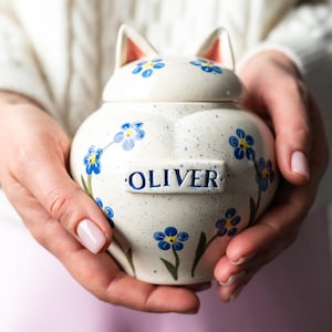 Könnte beinhalten: Cremefarbene Keramik-Urne in Katzenform mit Deckel, verziert mit blauen Vergissmeinnicht-Blumen. Die Urne trägt den Namen "OLIVER" in blauer Schrift. Die Katzenohren auf dem Deckel sind innen rosa.