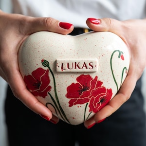 May include: Heart-shaped ceramic urn with red poppy floral design and the name "LUKAS" in a rectangular plaque. The urn is off-white with red speckles and green stems. The urn is held in front of a white shirt.