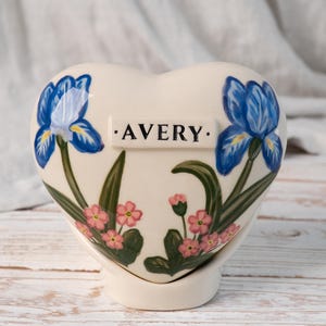 May include: A heart-shaped ceramic keepsake urn in cream with hand-painted blue iris flowers and small pink blossoms. The centre features a raised rectangular plaque with the name AVERY printed in black capital letters on a white background.
