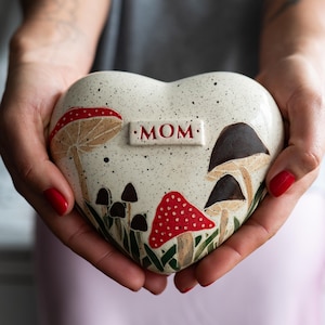 Könnte beinhalten: Herzförmiges Keramik-Andenken mit gesprenkeltem cremefarbenem Hintergrund, mit Pilzillustrationen in Rot, Braun und Schwarz. Das Wort "MOM" ist rot dargestellt. Das Andenken wird in zwei Händen gehalten.