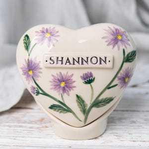 Può includere: Scatola portarricordi in ceramica a forma di cuore, decorata con fiori viola e foglie verdi. Il nome "SHANNON" è stampato su una placca rettangolare. La scatola è di colore crema e poggia su una base abbinata.