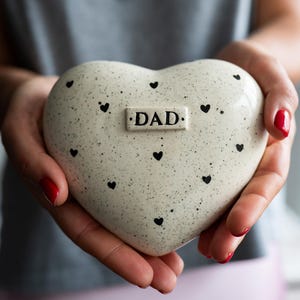May include: A cream-coloured, heart-shaped ceramic keepsake, speckled with black dots and small heart motifs. The word "DAD" is inscribed on a rectangular plaque. The keepsake is held in open hands.