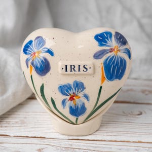 May include: Heart-shaped ceramic container with blue iris flower illustrations and the word "IRIS". The container is off-white with a speckled finish, and the flowers have yellow and orange accents. The container sits on a matching base.