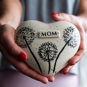 May include: Cream-coloured heart-shaped ceramic keepsake with a dandelion design and the word "MOM". The urn has black dandelion illustrations.
