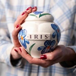 Può includere: Un'urna in ceramica bianca con coperchio, ornata da motivi floreali blu e la parola "IRIS" in lettere viola. L'urna è tenuta in mano con smalto rosso, su uno sfondo a quadri.