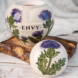 May include: A cream-colored ceramic jar with a matching lid, featuring hand-painted purple thistle flowers and green leaves. The jar has a rectangular plaque on the front with the word ENVY printed in black. The surface has small dark speckles.