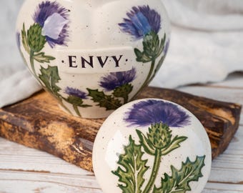 Puede incluir: Un tarro de cerámica color crema con tapa a juego, decorado con flores de cardo moradas y hojas verdes pintadas a mano. El tarro tiene una placa rectangular en el frente con la palabra ENVY impresa en negro. La superficie tiene motas oscuras.