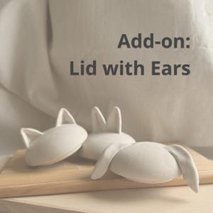 May include: Three white ceramic lids with ear-shaped handles are displayed on a wooden surface. The lids vary in ear design, with some upright and others drooping. Text on the image reads "Add-on: Lid with Ears."