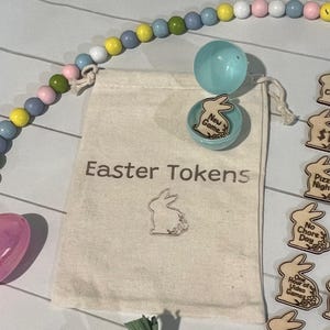 May include: A beige drawstring bag with the text "Easter Tokens" and a bunny graphic. A wooden bead garland in pastel colours and wooden tokens with bunny shapes are also visible. A blue plastic egg and a blue container with a wooden bunny are also present.