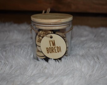 May include: A clear glass jar with a wooden lid and twine detailing. The jar is filled with small wooden discs. A round wooden tag with the words "I'M BORED!" is attached. The jar rests on a white, fluffy surface.