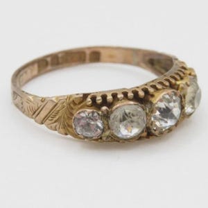 Könnte beinhalten: Ein goldfarbener Ring mit fünf klaren Edelsteinen. Der Ring hat ein verziertes Design mit geätzten floralen Mustern auf dem Band. Die Edelsteine sind in einer Reihe angeordnet.