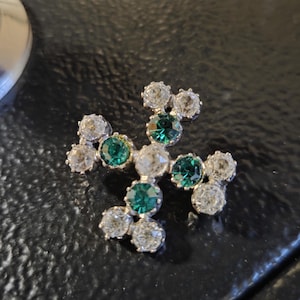 Puede incluir: Un broche en forma de cruz con una piedra central transparente rodeada de piedras redondas facetadas, alternando entre verde esmeralda y transparente, engastadas en un marco de metal plateado. Doce piedras forman un patrón de cruz simétrico.