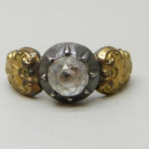 May include: A ring featuring a central round faceted clear stone set in a dark oxidized metal bezel with prongs. The band is composed of two gold-toned side sections, each displaying an embossed fan-like pattern with curved decorative details.