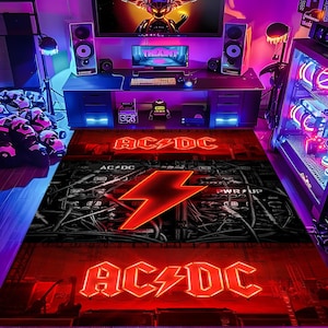 Puede incluir: Una alfombra rectangular con el logotipo de AC/DC en rojo y un rayo rojo. La alfombra tiene un fondo negro y gris con un diseño de cables y maquinaria. La alfombra está en una habitación con una configuración de computadora.