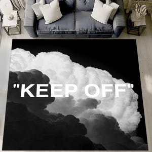Può includere: Tappeto rettangolare in bianco e nero con un design a nuvola e la scritta bianca "KEEP OFF". Il tappeto è posato su un pavimento in legno, con un divano e cuscini sullo sfondo.