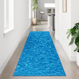 May include: A rectangular blue rug with a water ripple design. The rug is placed in a hallway with white walls and a concrete floor. The rug's pattern mimics the look of sunlight reflecting on water.