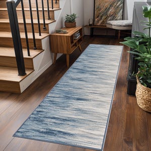 May include: A long rectangular runner rug featuring a horizontal abstract pattern in shades of navy blue, light blue, and white. The rug has a thin dark blue border and is positioned on a dark wood floor in a hallway with stairs and furniture.