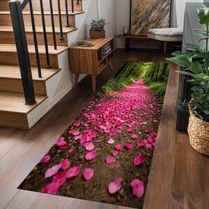 Puede incluir: Una alfombra rectangular para interiores que representa un camino forestal cubierto de pétalos de flores rosas. El diseño crea una ilusión de profundidad, con el camino que conduce a un dosel verde de árboles. La alfombra está colocada sobre un suelo de madera.