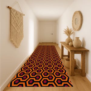 May include: A hallway runner rug with a repeating geometric pattern in orange, black, and burgundy. The rug is placed in a hallway with white walls, a wooden bench, and a decorative wall hanging.