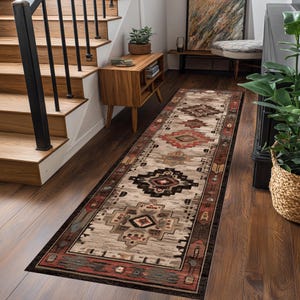 Puede incluir: Una alfombra rectangular larga con un patrón geométrico en tonos beige, marrón, rojo y negro. La alfombra está colocada sobre un suelo de madera en un pasillo. En el fondo se ven una escalera y muebles de madera. La alfombra mide unos 240 cm de largo.