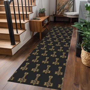 Può includere: Un tappeto runner nero con un motivo ripetuto di simboli dorati del martello di Thor. Il tappeto è rettangolare e posizionato su un pavimento in legno, aggiungendo un elemento decorativo all'ingresso. Misura circa 200 cm di lunghezza.