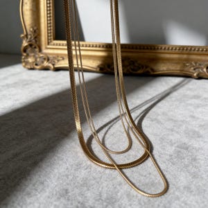 May include: A multi-strand gold-toned necklace featuring three distinct chains with a flat, herringbone-style link pattern. The chains hang in cascading loops, displaying a metallic sheen and a smooth, flexible texture that catches the light.
