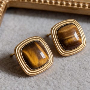 May include: A pair of square stud earrings with a gold-colored frame. Each earring features a polished, square tiger's eye stone with brown and gold tones. The earrings are set against a neutral gray background.
