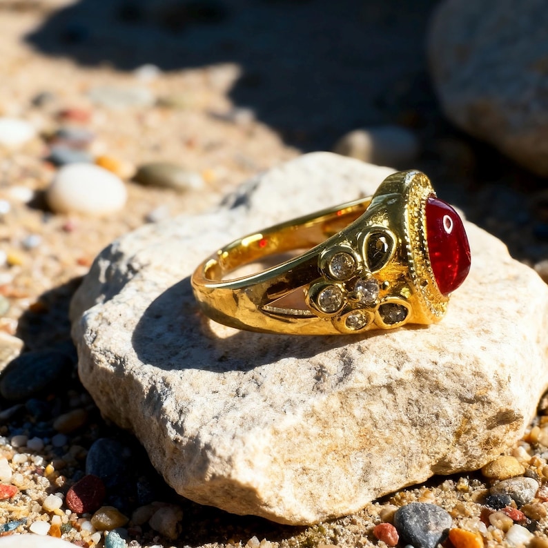 May include: A gold ring with a large, oval, red gemstone and small, clear accent stones. The ring is set on a light-colored rock, with a sandy background. The ring's band has intricate detailing.