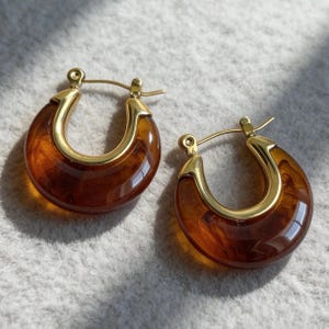 May include: A pair of hoop earrings featuring a polished gold-tone metal frame that secures a semi-circular, translucent amber-colored resin piece. The design includes a hinged post closure mechanism, showcasing a smooth, glossy finish on both the metal and the resin components.
