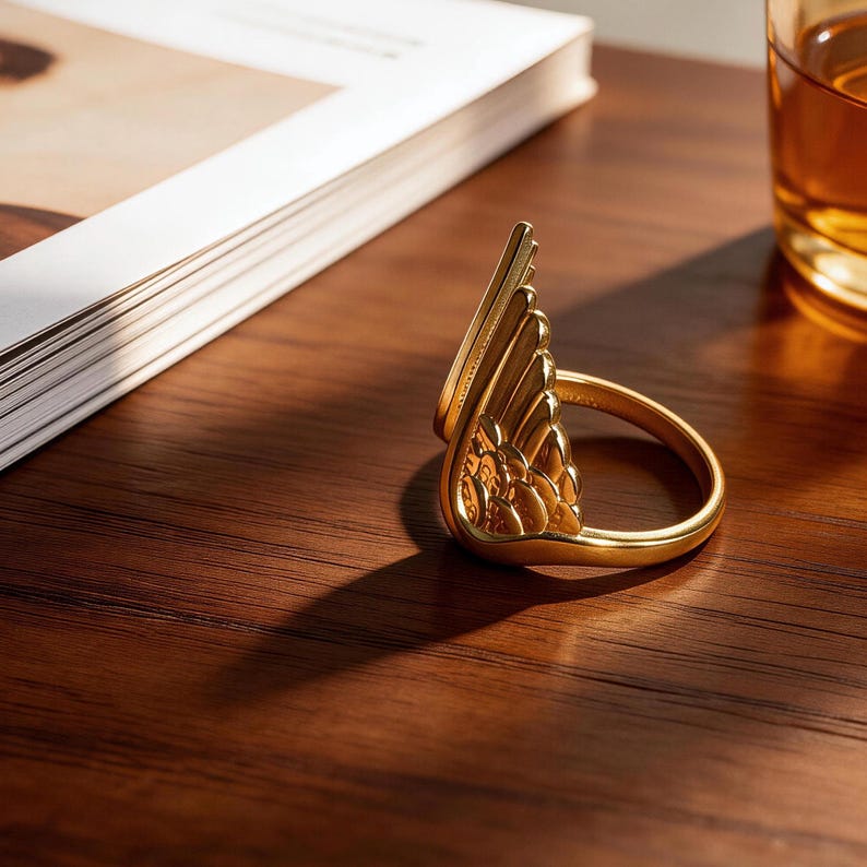 Może przedstawiać: Złoty pierścionek ze skrzydłem, leżący na drewnianej powierzchni. Pierścionek ma szczeg&oacute;łowe elementy przypominające pi&oacute;ra i znajduje się obok książki i szklanki płynu.