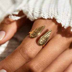 Może przedstawiać: Złoty pierścionek z motywem skrzydeł. Pierścionek posiada dwa otwarte skrzydła, każde ze szczeg&oacute;łowymi wzorami pi&oacute;r. Pierścionek jest noszony na palcu, prezentując skomplikowany design i ciepły, metaliczny odcień biżuterii.
