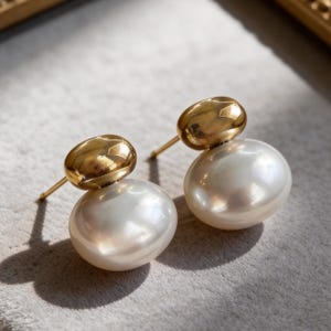 May include: A pair of gold stud earrings. Each earring has a large, lustrous white pearl base with a smaller, oval gold accent. The earrings are set against a soft, neutral background.