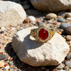 May include: A gold ring with a large, oval, red gemstone at the center, surrounded by smaller clear stones. The ring is set on a light-colored rock, with a background of other rocks and sand.
