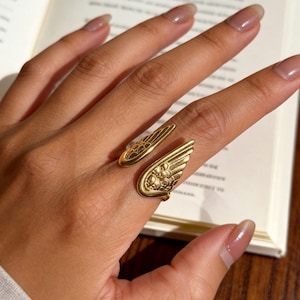 Może przedstawiać: Złoty pierścionek z motywem skrzydeł. Pierścionek owija się wok&oacute;ł palca z dwoma szczeg&oacute;łowymi kształtami skrzydeł. Skrzydła mają skomplikowane wzory i tekstury, tworząc delikatny i elegancki wygląd. Pierścionek jest noszony na dłoni.