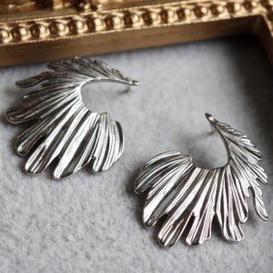 May include: A pair of silver-toned earrings featuring a fan-like, organic shape with deep, vertical ridges and a polished, reflective finish. The earrings have a curved, semi-circular silhouette that mimics the appearance of a stylized leaf or fin.