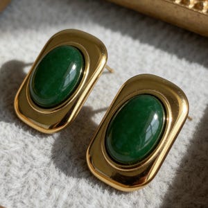 May include: A pair of gold-coloured stud earrings. Each earring has a rectangular gold frame with a polished, oval-shaped, dark green stone centre. The earrings are displayed on a light grey, textured surface.