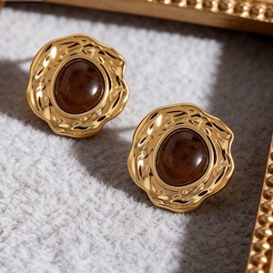 May include: A pair of stud earrings featuring a central smooth, dark brown circular stone set within a textured, irregular gold-toned metal frame. The metal surface has a hammered, wavy appearance that surrounds the polished brown center piece.