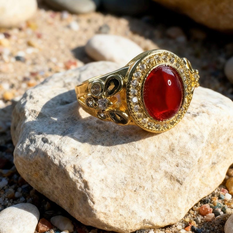 May include: A gold-colored ring with a large, oval, red gemstone at the center. The ring is set with small, clear stones around the red stone and on the band, which features floral designs.