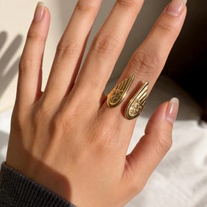 Może przedstawiać: Złoty pierścionek z motywem skrzydeł. Pierścionek ma dwa otwarte skrzydła, każde ze szczeg&oacute;łowymi wzorami pi&oacute;r. Pierścionek jest noszony na palcu, prezentując skomplikowany design i metaliczny połysk. To biżuteria.