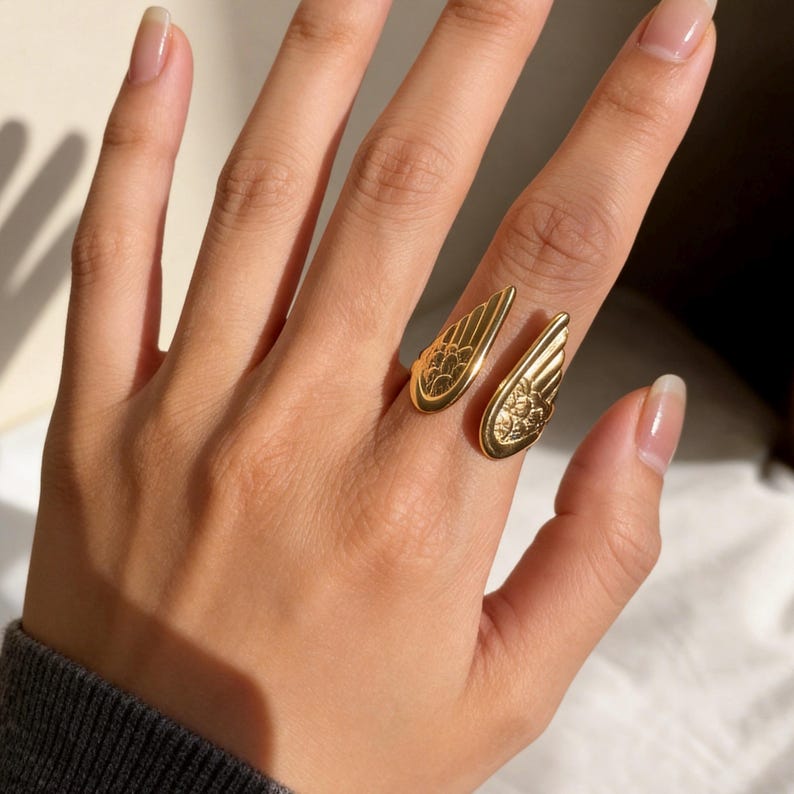 Może przedstawiać: Złoty pierścionek z motywem skrzydeł. Pierścionek ma dwa otwarte skrzydła, każde ze szczeg&oacute;łowymi wzorami pi&oacute;r. Pierścionek jest noszony na palcu, prezentując skomplikowany design i metaliczny połysk. To biżuteria.