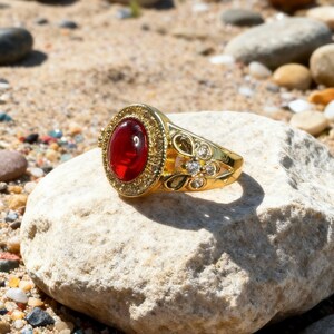 May include: A gold-colored ring with a large, oval, red gemstone. The ring has a detailed design with small clear stones and floral accents. The ring is displayed on a light-colored rock, with a sandy background.