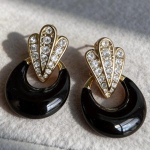 May include: A pair of gold-tone earrings featuring a fan-shaped top section encrusted with clear round rhinestones. Attached to the base of each fan is a smooth, glossy black circular hoop. The earrings have a post-back closure mechanism.