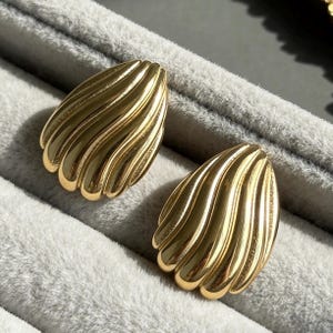 Peut inclure: Une paire de boucles d'oreilles dorées présentant un design en forme de goutte d'eau polie avec des crêtes verticales en relief créant un aspect de coquillage. Les boucles d'oreilles ont une surface lisse et réfléchissante.