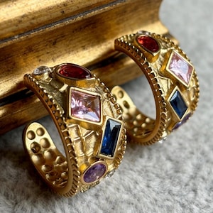 May include: A pair of gold-toned hoop earrings with a textured, hammered surface. Each hoop is set with multi-colored, multi-shaped faceted crystals, including red, pink, blue, and purple stones arranged in a scattered, geometric pattern.