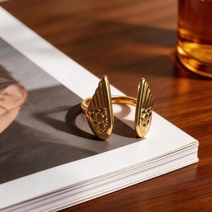 Może przedstawiać: Złoty pierścionek o otwartym designie, z dwoma stylizowanymi elementami w kształcie skrzydeł. Pierścionek umieszczony jest na białej powierzchni z rozmytym tłem, ukazującym szczeg&oacute;ły i design biżuterii.