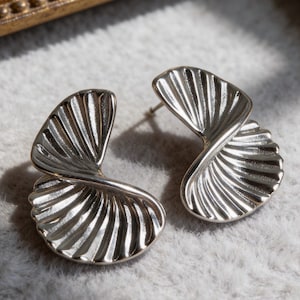 May include: A pair of silver-toned stud earrings featuring a fan-like, pleated shell design. Each earring has a curved, semi-circular shape with radiating ridges that create a textured, metallic surface. The earrings are secured with a post back.