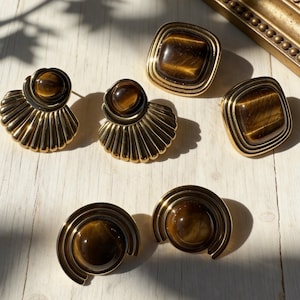 May include: A collection of six gold-tone metal earrings featuring polished tiger's eye gemstones. The set includes two fan-shaped earrings, two square-shaped earrings with rounded corners, and two circular earrings with concentric semi-circular metal detailing.