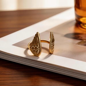 Może przedstawiać: Złoty pierścionek ze skrzydłem. Pierścionek ma dwa stylizowane skrzydła, po jednym z każdej strony, połączone obrączką. Skrzydła mają szczeg&oacute;łowe wzory pi&oacute;r. Pierścionek leży na białej i beżowej powierzchni.