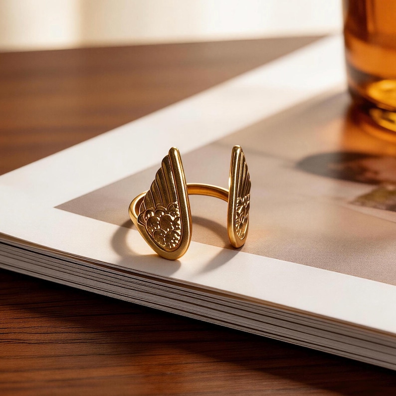 Może przedstawiać: Złoty pierścionek ze skrzydłem. Pierścionek ma dwa stylizowane skrzydła, po jednym z każdej strony, połączone obrączką. Skrzydła mają szczeg&oacute;łowe wzory pi&oacute;r. Pierścionek leży na białej i beżowej powierzchni.